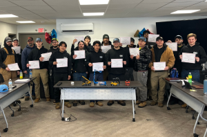 A group of people stand in a classroom holding certificates, posing for a photo in front of tables with model construction equipment and drinks.