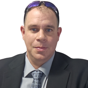A man wearing a black suit, gray shirt, patterned tie, and sunglasses resting on his head, posing in front of a white background.