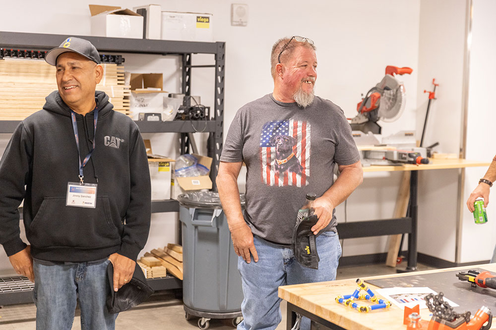 Two men stand and smile in a workshop, one wearing a gray t-shirt with an American flag design, the other in a black hoodie and cap. Tools and equipment are visible on tables and shelves around them.